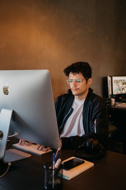 Focused young man with eyeglasses working at a desktop computer in a modern office setting.