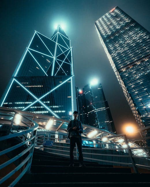 A person stands under vibrant skyscrapers in Hong Kong at night, featuring bright neon lighting.