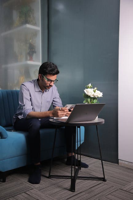 Concentrated young man using laptop indoors, seated on blue couch with smartphone.