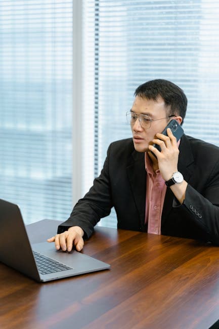 Professional man engaging in a phone call while working on a laptop at an office table.