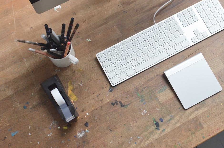 From above of artist workplace with modern keyboard and stationary placed on wooden desk stained with paint